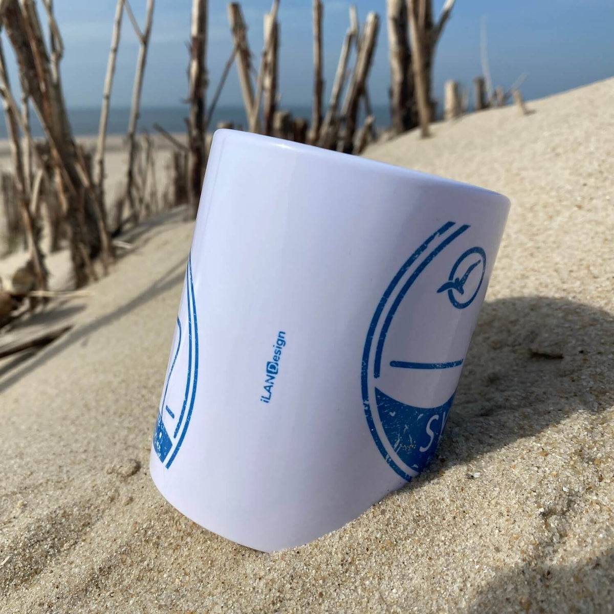 Weißer Becher liegt am Strand auf Sylt. Im Hintergrund ist das blaue Meer zu sehen. Der Becher ist in blau bedruckt mit dem Begriff Sylt, einem Strandkorb sowie einer Möwe die durch die Sonne fliegt.