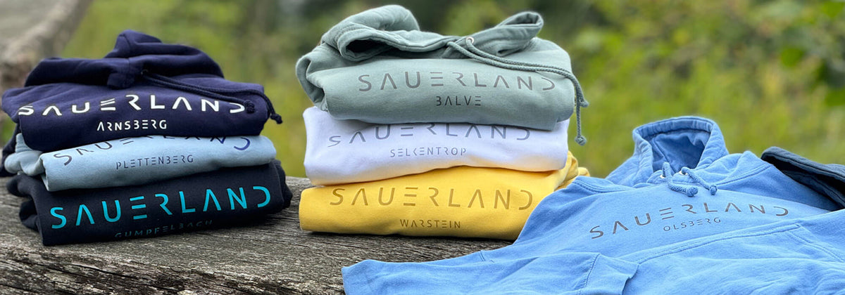 Sauerland Hoodies mit Dorfnamen liegen zusammengefaltet auf einer Holzbank im Grünen. Die Hoodiefarben reichen von verschiedenen Blautönen, über grün-grau, gelb bis hi zu klassischem weiß. Unter dem modernen Sauerland-Schriftzug wird ein individueller Dorfname iengedruckt.