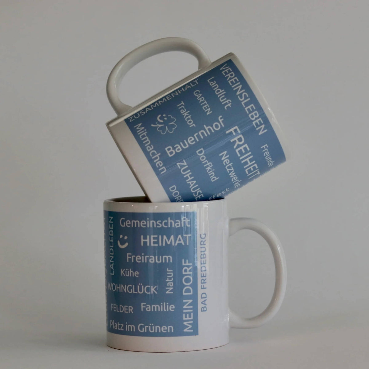Heimatbecher aus weißer Keramik mit blauer Banderole und typischen Heimatbegriffen als Aufdruck. Mit Dorfnamen individualisierbar. 
