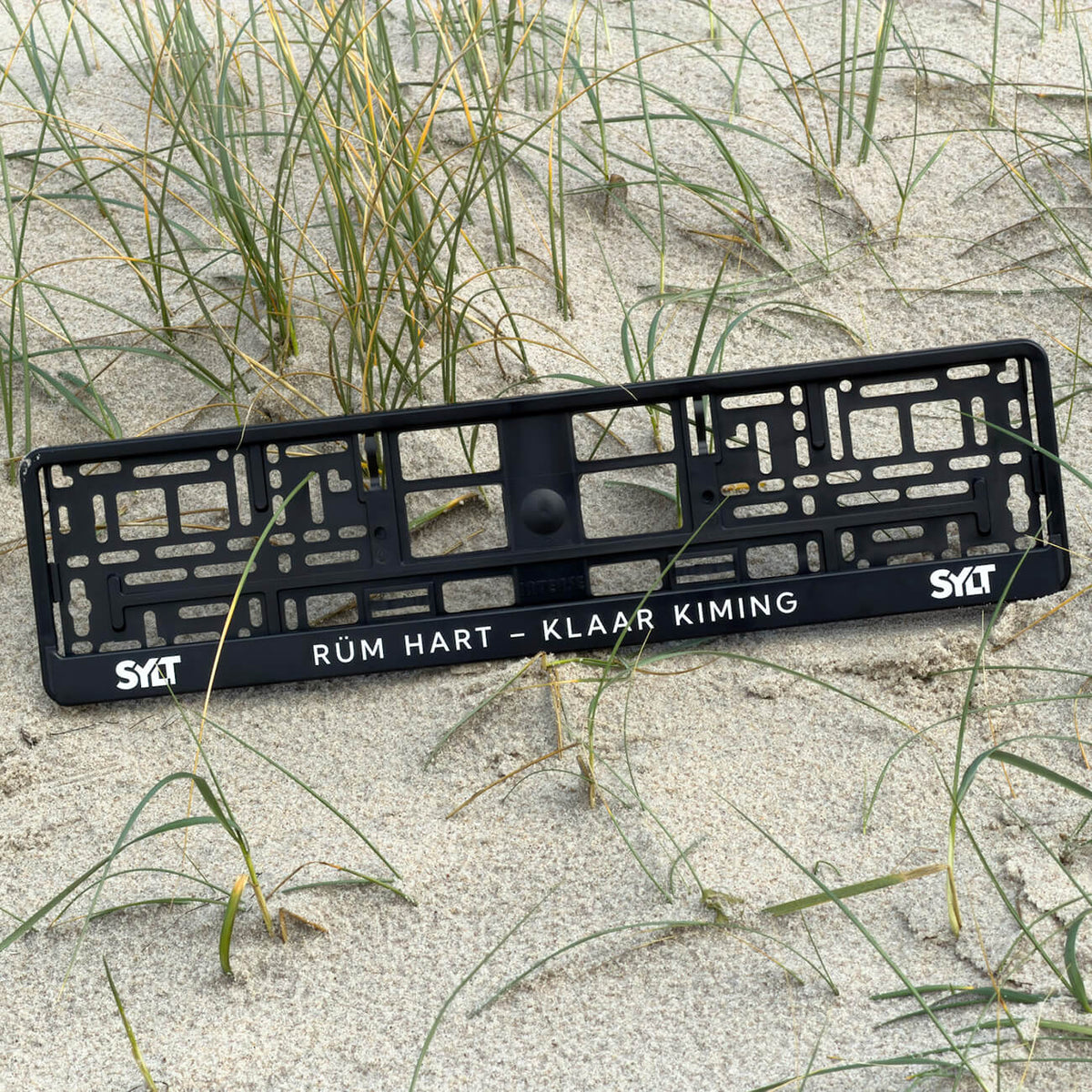 Sylt Kennzeichenhalter mit modernem Sylt-Schriftzug links und rechts. In der Mitte steht der friesische Spruch "Rüm hart – Klaar Kiming". Der Nummernschildhalter liegt im Sand zwischen kleinen Dünengrasbüscheln.