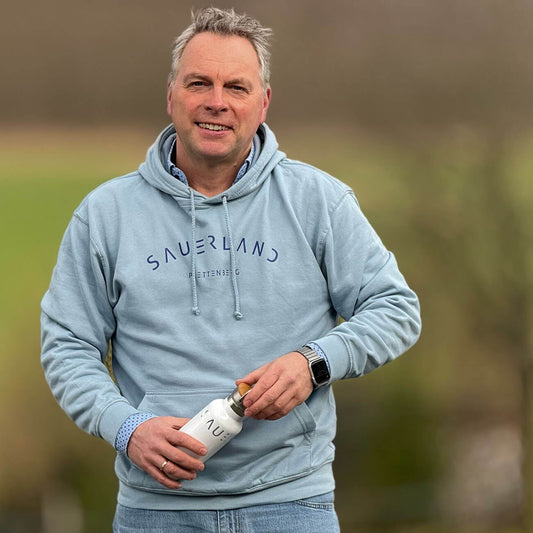 Mann trägt einen hellblauen Hoodie mit dem modernen Sauerland Schriftzug leicht gebogen. Darunter steht ebenfalls in dunkelblau der Dorfname Plettenberg